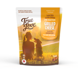 Bag of True Love dog treats with a woman smiling at her dog on the grass. Grilled Cheese flavour - golden cheddar and vine-ripened tomato.